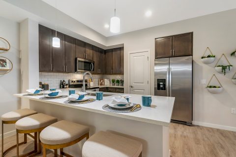 Model apartment kitchen with dark wood cabinets, subway tile backsplash, quartz countertops, stainless steel appliances, and barstool seating at counter