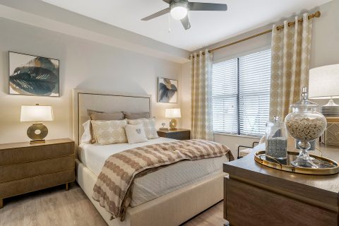 Model apartment bedroom with queen bed, end tables, large window, and ceiling fan
