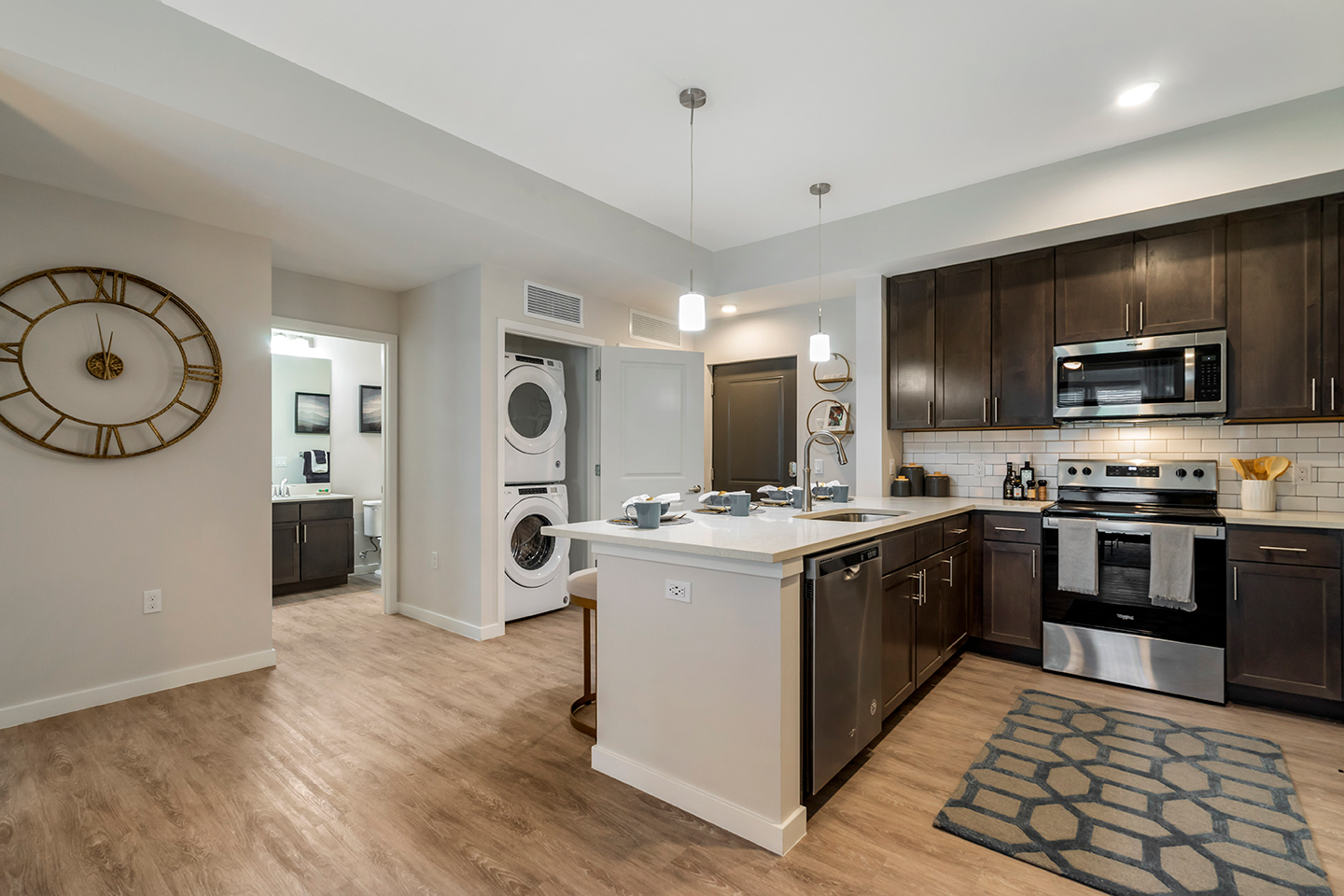 Model apartment kitchen with dark wood cabinets, wood-style plank flooring, and stainless steel appliances with view of washer and dryer machines